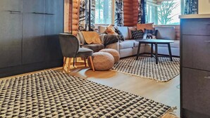 A cozy living room with a sofa, armchairs, and a coffee table. There are patterned rugs on the floor and a wooden ceiling.