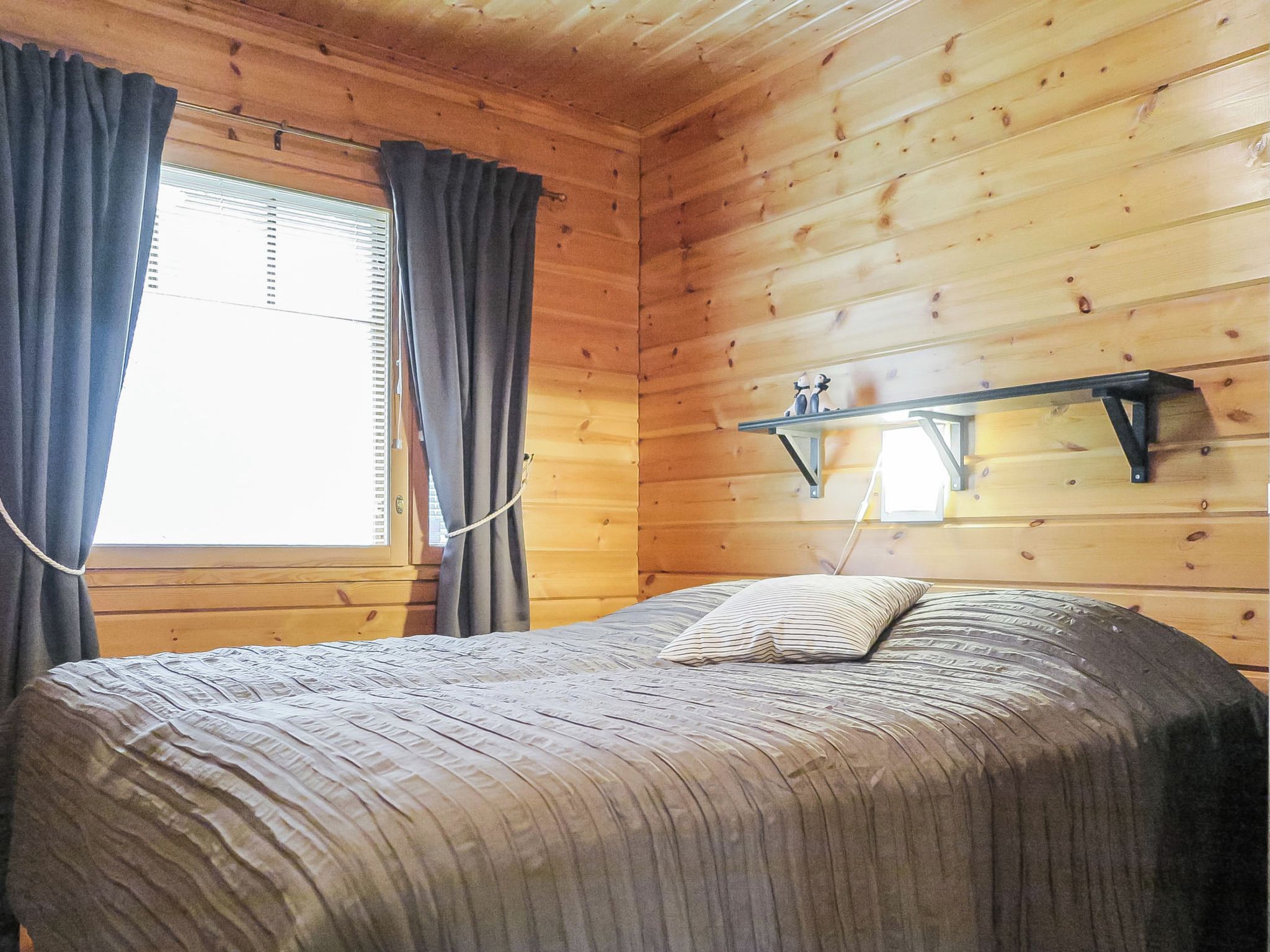 A wooden room with a bed, a window with curtains, and a shelf with items.