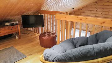 An attic room with a wooden ceiling, a television on a wooden stand, a brown leather ottoman, a grey cushioned chair, and a wooden railing.