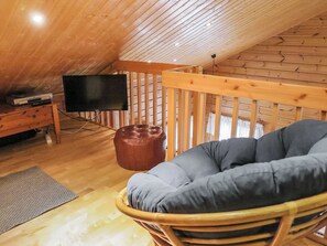 An attic room with a wooden ceiling, a television on a wooden stand, a brown leather ottoman, a grey cushioned chair, and a wooden railing. - Artturi by Interhome (Kittilä)