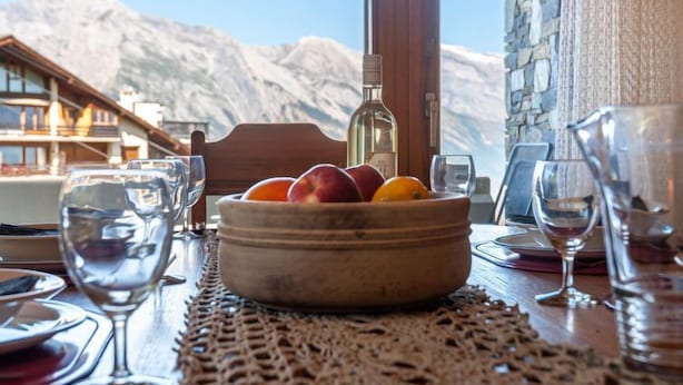 Un tavolo con una ciotola di frutta, una bottiglia di vino e bicchieri, con vista sulle montagne.