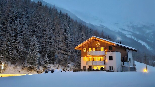 Een houten chalet met twee verdiepingen, met een ster op het dak, omringd door met sneeuw bedekte bomen en bergen.