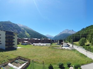 Un quartier résidentiel où se côtoient des bâtiments modernes et traditionnels, avec un terrain herbeux et des montagnes en toile de fond.