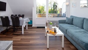 Moderni olohuone, jossa on sohva, ruokapöytä ja tuoleja. Suuri ikkuna avautuu ulos ja tarjoaa näkymän ulkoilumaastoon.