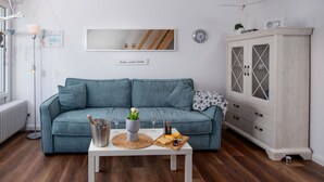 Olohuoneessa on sininen sohva, valkoinen sohvapöytä, jonka päällä on samppanjapullo, lasit ja juustolautanen. Huoneessa on myös valkoinen kaappi, jossa on lasiovet, ja seinällä on kello.