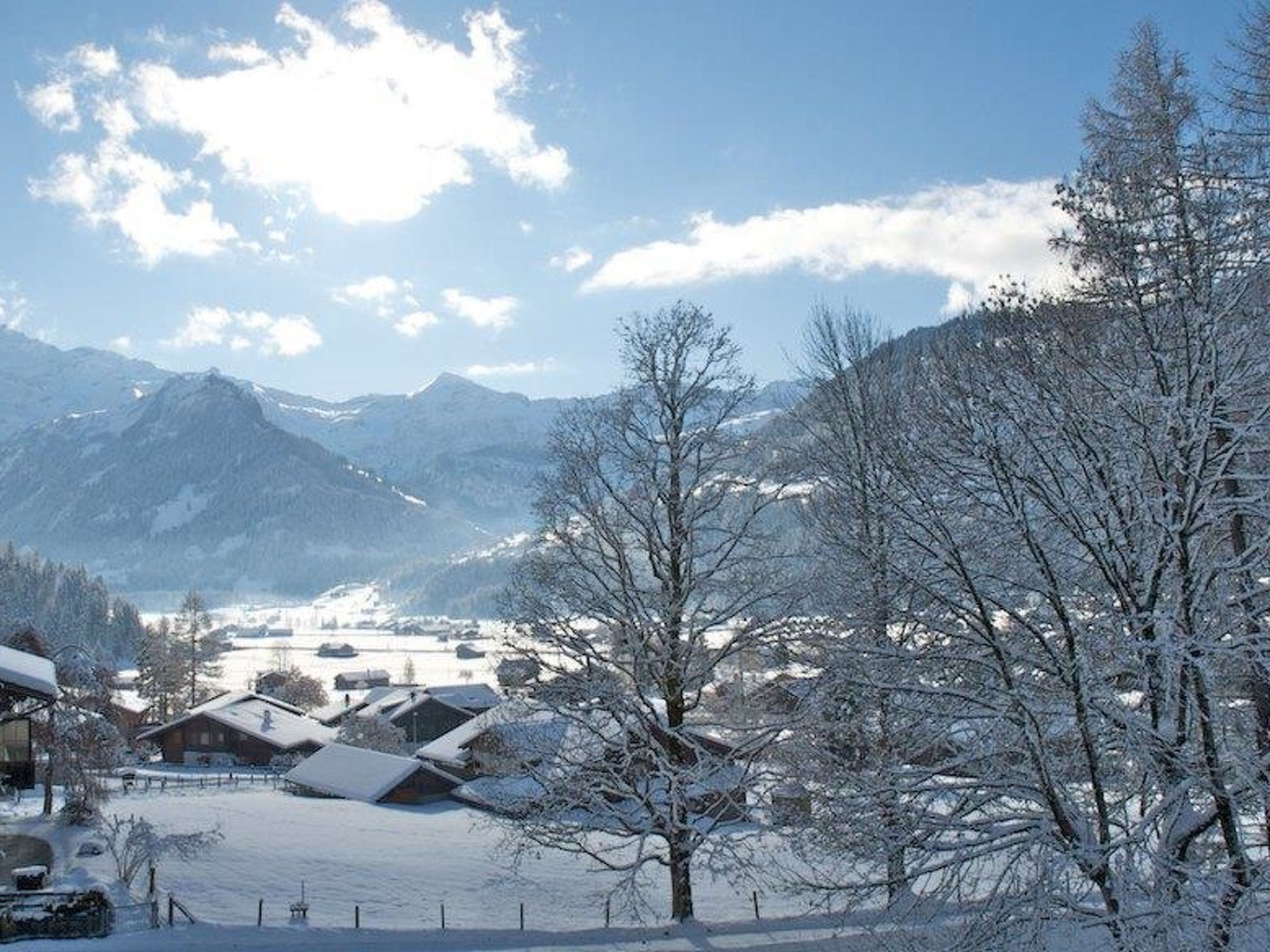 A snowy landscape with mountains in the background, a few buildings, and leafless trees.