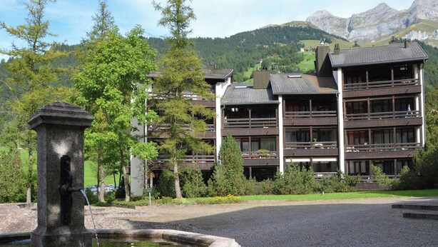 Un edificio con diversi balconi, una fontana in pietra e montagne sullo sfondo.