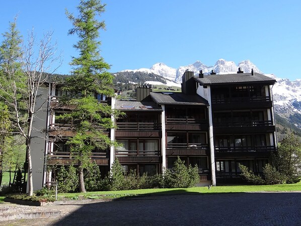 A multi-story building with a modern architectural design, featuring balconies and large windows, set against a backdrop of snow-capped mountains.
