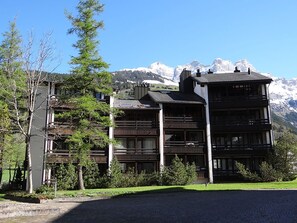 Un immeuble à plusieurs étages doté de balcons, entouré d’arbres et avec une chaîne de montagnes en arrière-plan.