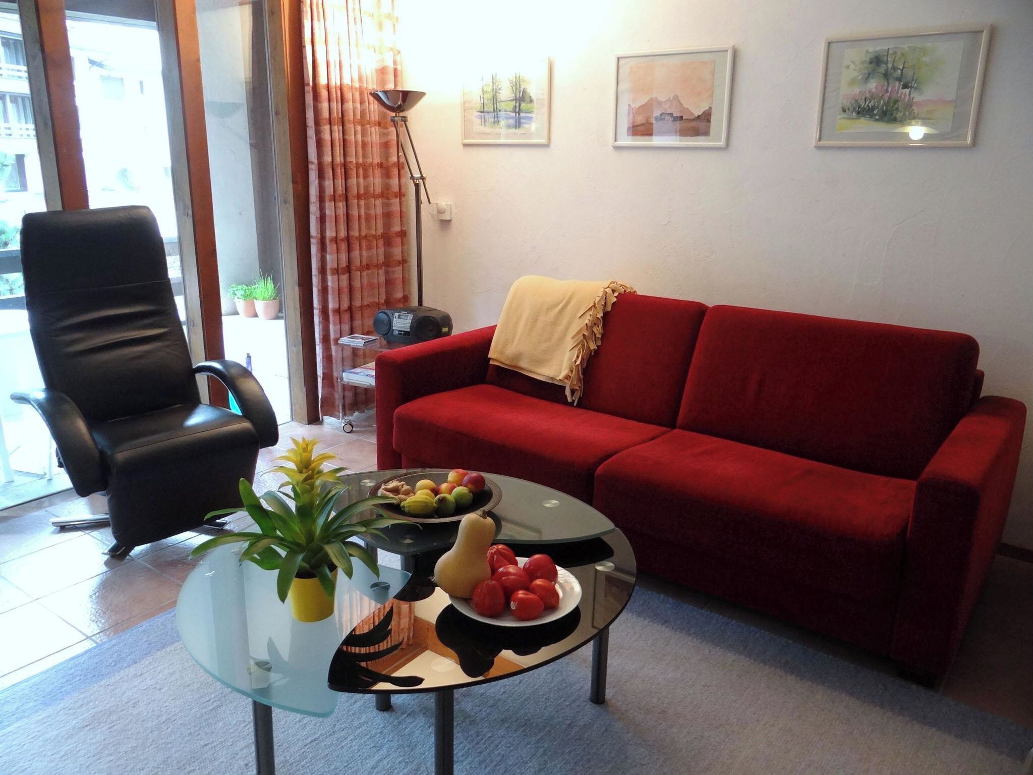 Una sala de estar con un sofá rojo, una silla negra, una mesa de centro de vidrio con fruta y una planta, y una lámpara.