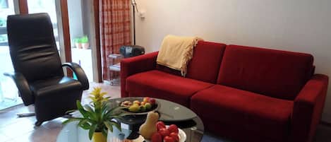 Una sala de estar con un sofá rojo, una silla negra, una mesa de centro de vidrio con fruta y una planta, y una lámpara.