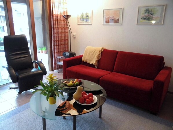 Un salon avec un canapé rouge, un fauteuil noir, une table basse en verre sur laquelle se trouvent des fruits et une plante, et une lampe.