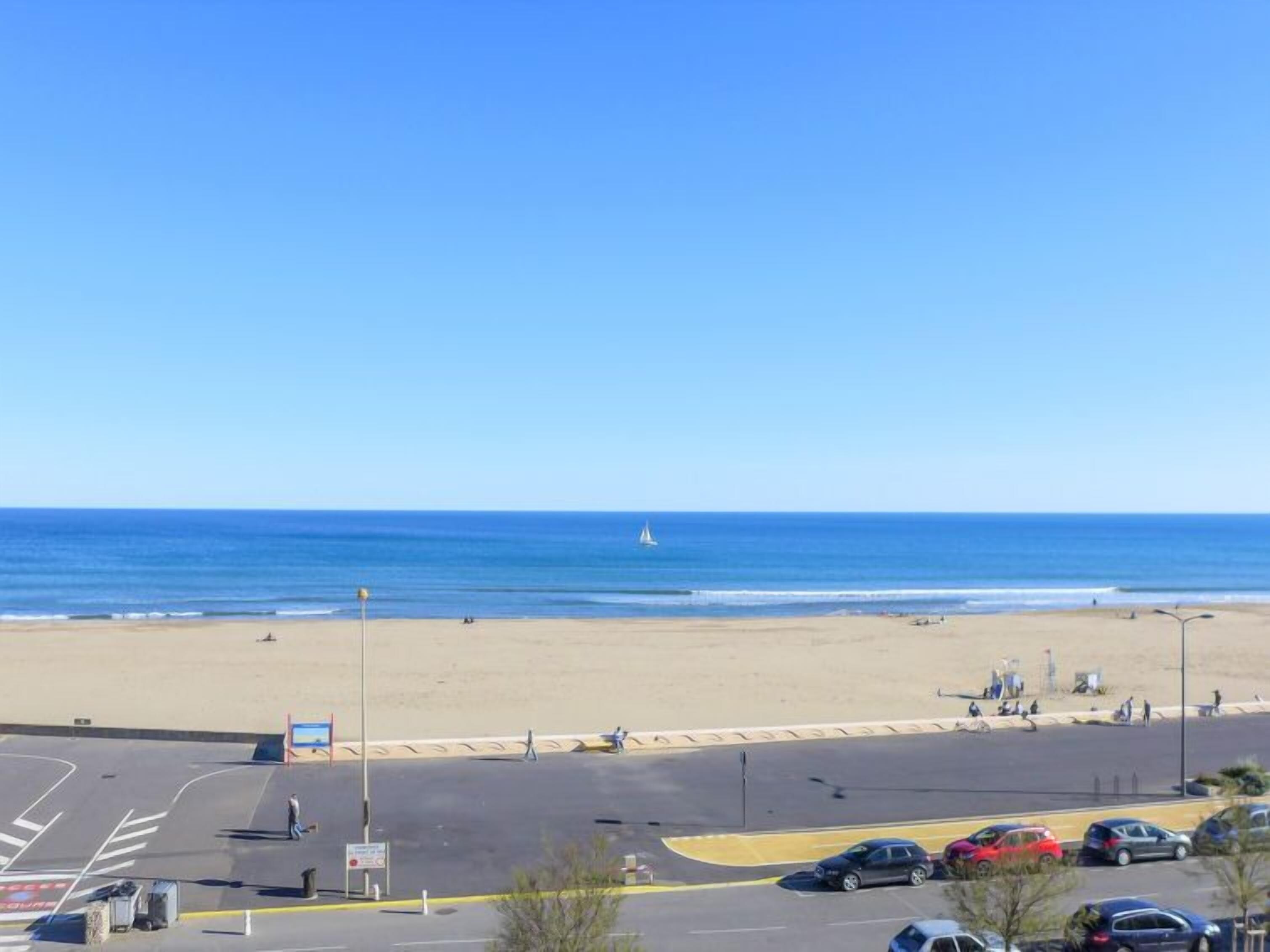 Une plage d’où l’on a une vue dégagée sur l’océan, avec un voilier qui se profile à l’horizon.