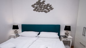 Cama doble con ropa de cama blanca y cabecera color turquesa, dos mesitas de noche con lámparas y un adorno en la pared.