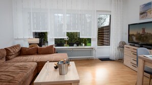 Una sala de estar con un sofá marrón, una mesa de centro de madera, un televisor y una ventana que ofrece vistas a zonas verdes y una maqueta de una casa.