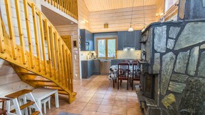 A wooden staircase leading to a second floor, a kitchen with dark cabinets and a stone fireplace, and a dining area with a table and chairs.