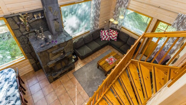 A cozy cabin interior with a stone fireplace, a wooden staircase, and large windows offering a view of the outdoors.