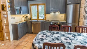 A compact kitchen with dark cabinets, a stainless steel refrigerator, a microwave, and a coffee maker. There is a window with blinds, a sink, and a dining area with a table and chairs.