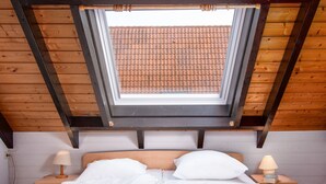 Een slaapkamer met dakraam, houten balken, een bed met witte lakens, twee nachtkastjes met lampen en uitzicht op een pannendak.