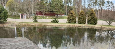 Eine Holzstege ragt in einen ruhigen See, wobei Bäume und ein rotes Gebäude im Hintergrund sich spiegeln.