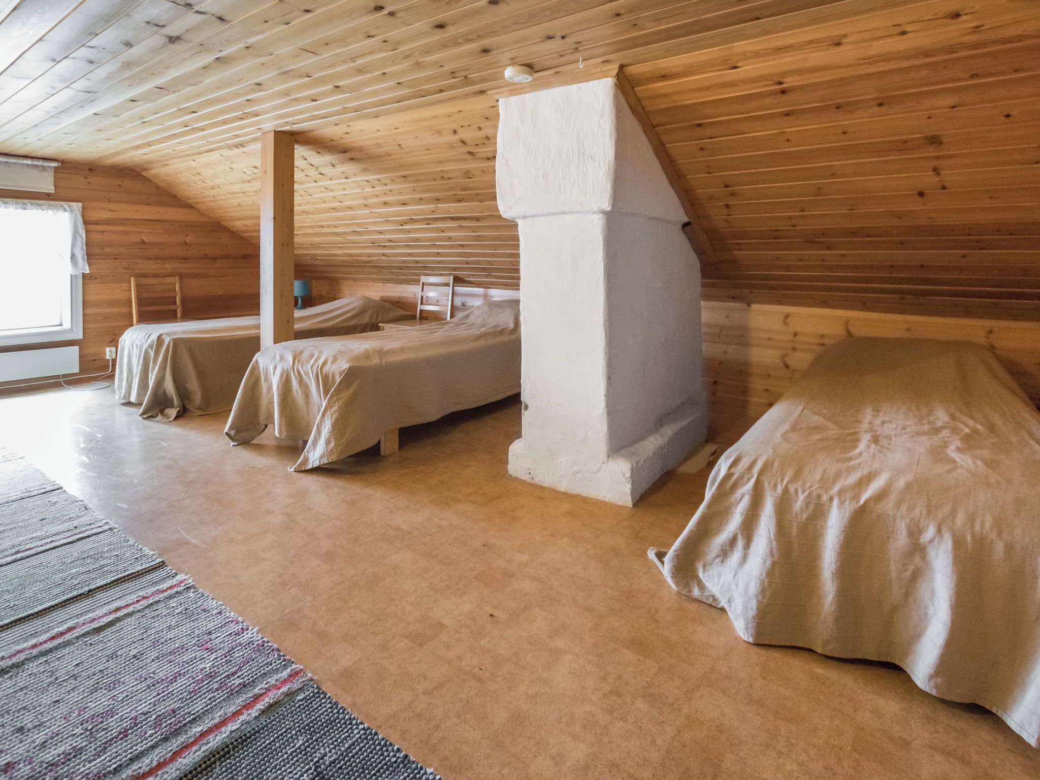 A room with a sloped ceiling, wooden walls, and a large window. There are two beds with white bedding and a rug on the floor.