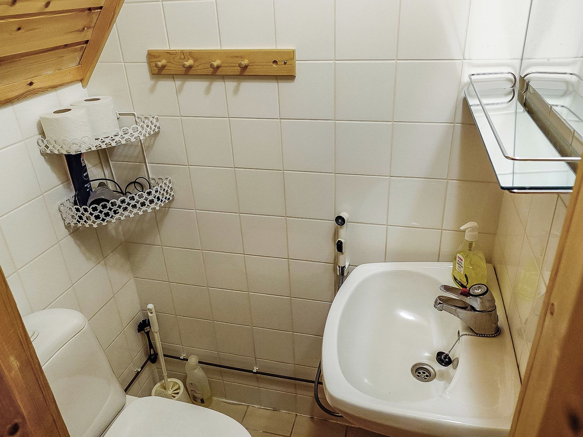 Een compacte badkamer met toilet, een wastafel en een douchehoek. Er zijn planken met toiletartikelen en een houten rek boven het toilet.