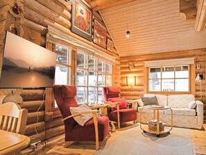 A cozy log cabin interior with a large TV, multiple seating areas, and large windows.