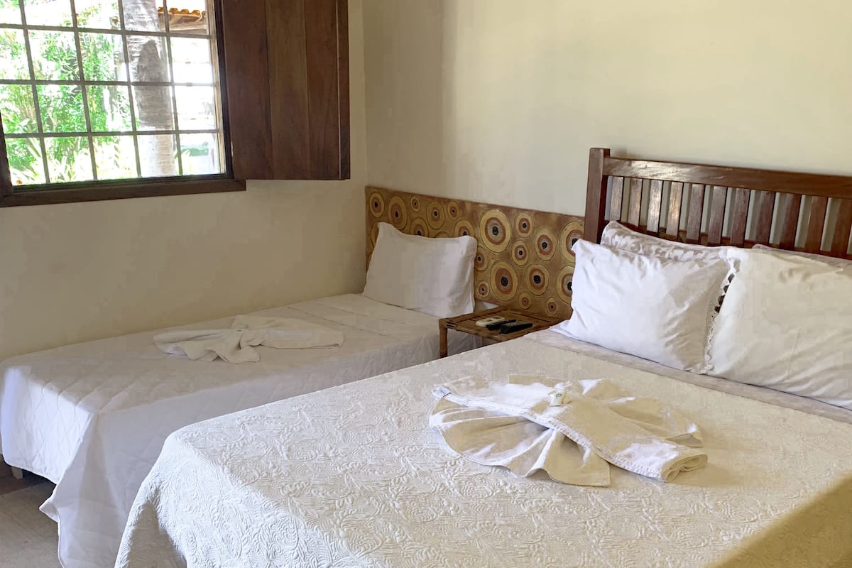 Habitación de hotel con dos camas individuales, cabecero de madera, una mesita pequeña y una ventana con vista a vegetación.