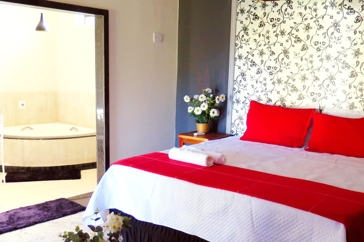 Un dormitorio con cama, una mesita con un florero, un cojín rojo y un papel tapiz con motivos florales.