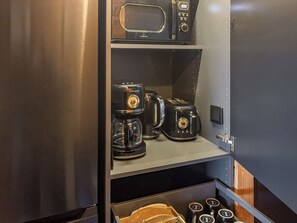 Une petite cuisine équipée d’une machine à café, d’un grille-pain et d’une bouilloire, le tout rangé sur des étagères.