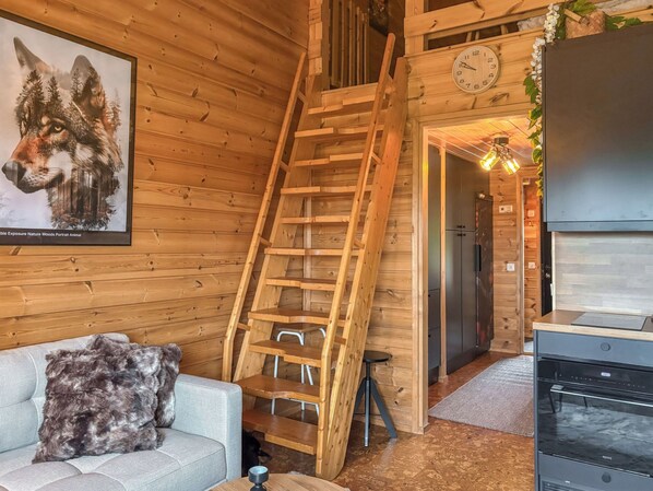 A wooden interior with a staircase, a sofa with a fur pillow, a framed animal portrait, and a kitchen area with appliances.