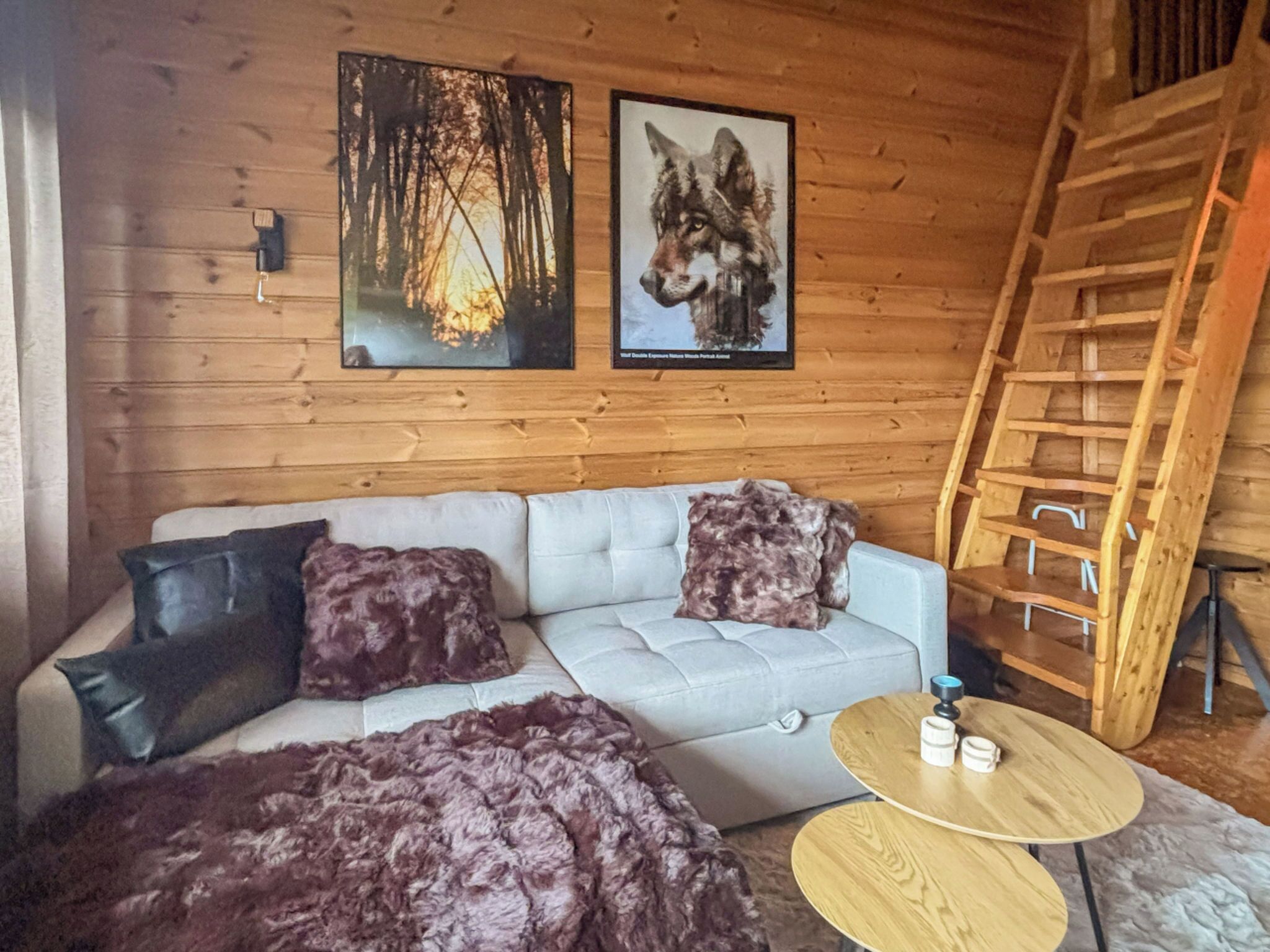 A cozy cabin interior with a sofa, two framed pictures, a wooden ladder, and a round wooden table.