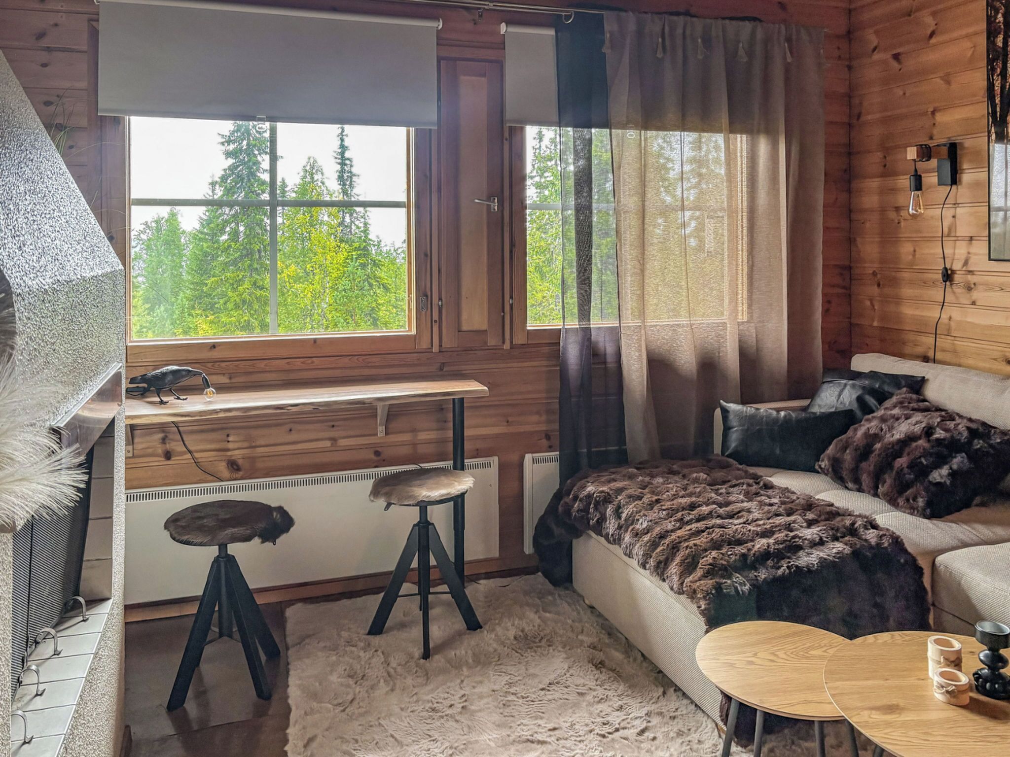 A cozy cabin interior with a wooden desk, a sofa, a round table, and a view of the forest through the windows.