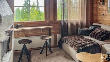 A cozy cabin interior with a wooden desk, a sofa, a round table, and a view of the forest through the windows.