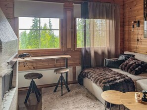Interior acogedor de una cabaña con un escritorio de madera, un sofá, una mesa redonda y vistas al bosque a través de las ventanas.