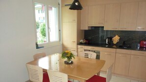 A dining area with a wooden table and chairs, a vase with flowers, and a kitchen with cabinets and appliances. - Blumenweg 10 Appartement/Fewo 6-Bettwohnung by Interhome (Engelberg)