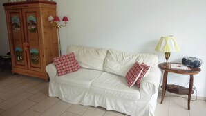 A living room with a white sofa, red and white checkered pillows, a wooden cabinet with decorative items, a floor lamp, and a small table with a radio. - Blumenweg 10 Appartement/Fewo 6-Bettwohnung by Interhome (Engelberg)