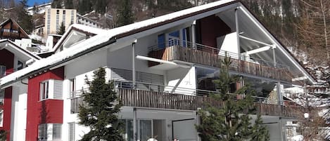 A two-story chalet with a balcony, surrounded by snow-covered trees and a mountainous backdrop.