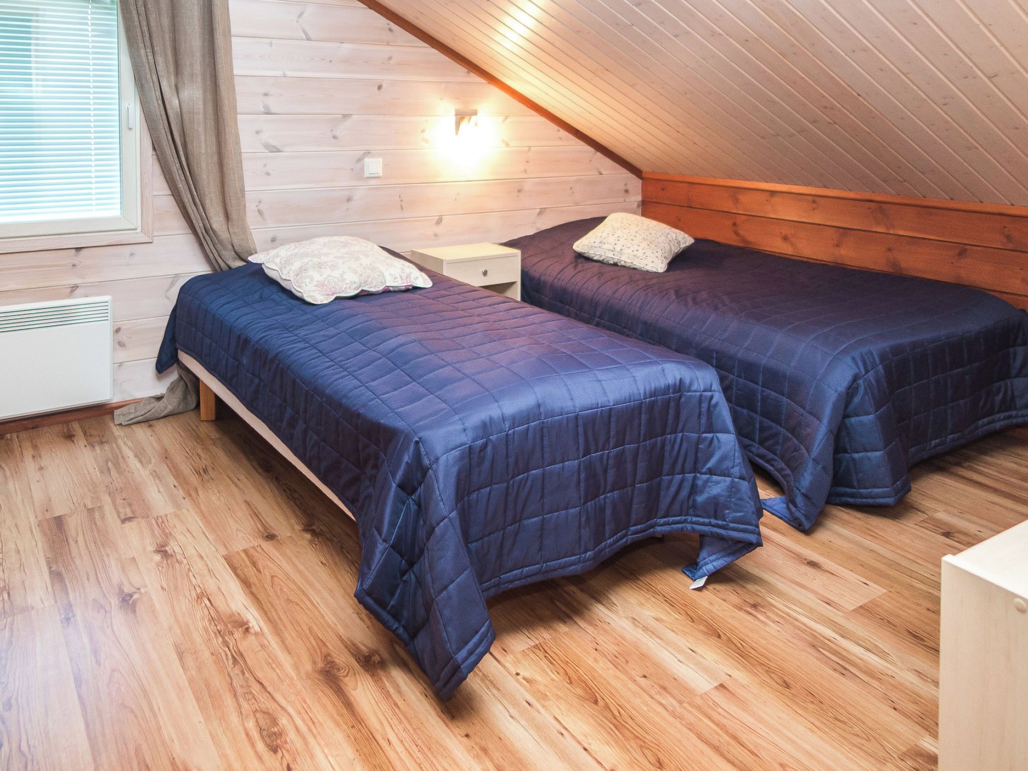 Ein Zimmer mit zwei Betten, Holzwänden, einer geneigten Decke und einem Fenster mit Jalousien.