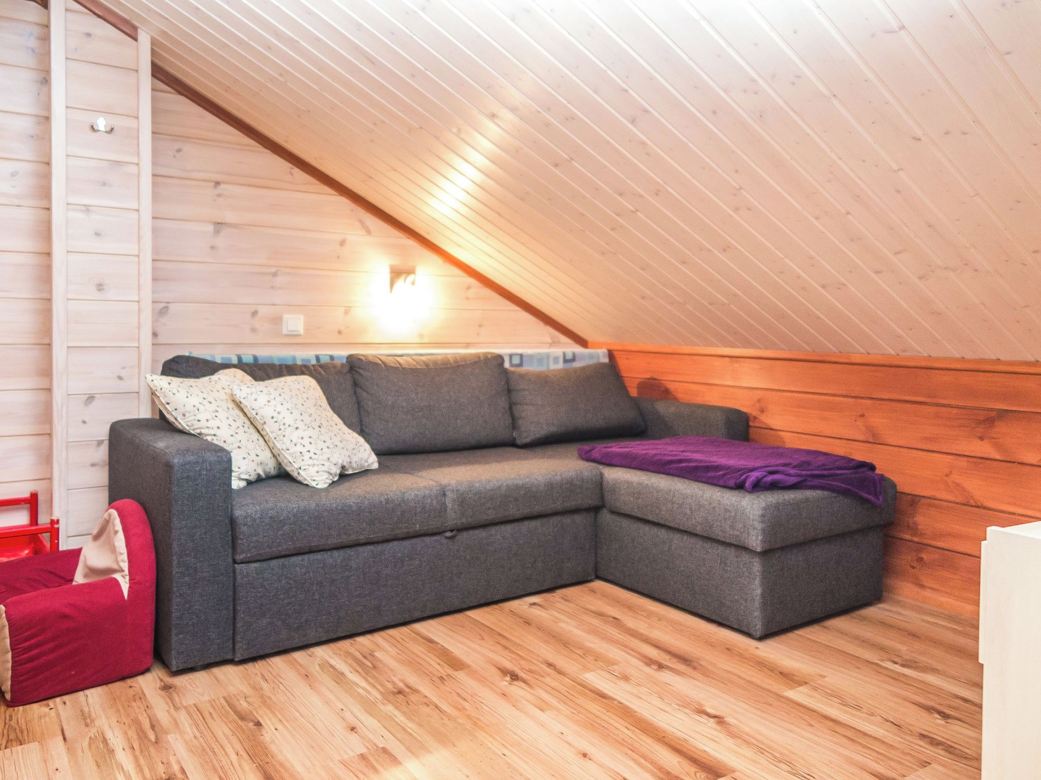 A cozy, single-level room with a wooden floor, a grey sectional sofa with pillows, a red chair, and a small red table.
