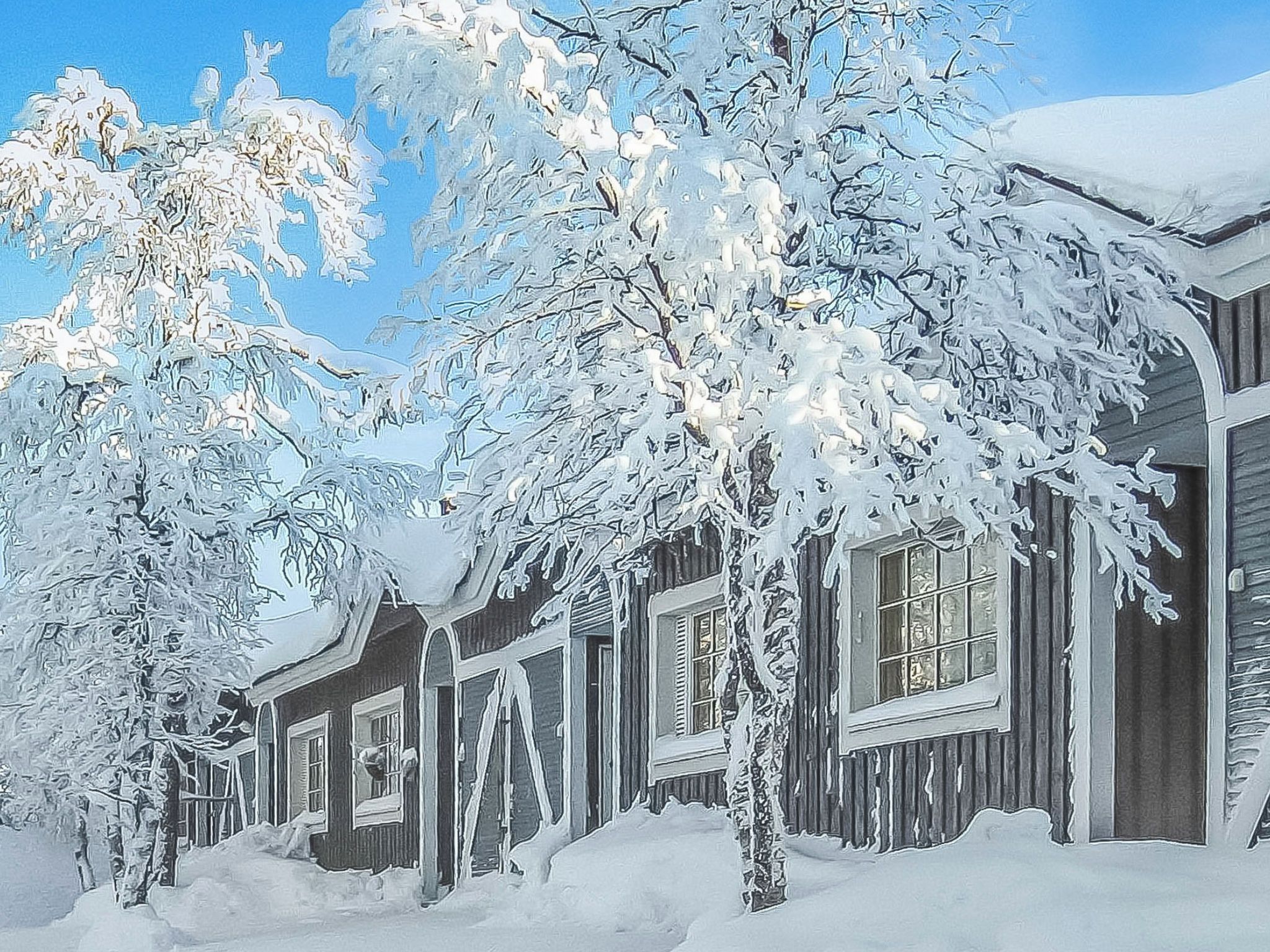 A row of wooden cabins with snow-covered roofs and trees, under a clear sky.
