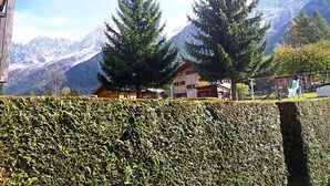 Un paisaje montañoso con dos árboles de hoja perenne destacados, un edificio de madera y un seto en primer plano.