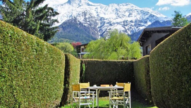 Un jardín con una mesa y sillas, rodeado de setos bien cuidados, y una cordillera en el fondo.