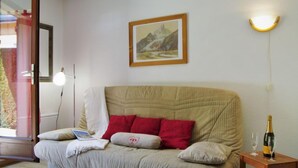 Una habitación acogedora con un sofá color beige, almohadas rojas, un cuadro enmarcado de un paisaje, una mesita de noche con una botella y vasos, y un espejo que refleja un interior colorido.