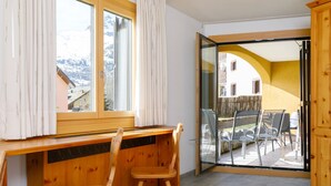 A room with a wooden desk and two chairs, a large window with curtains, and a sliding glass door leading to an outdoor area with chairs and a table. - Residenza Chesa Margun 23-5 by Interhome (Silvaplana-Surlej)