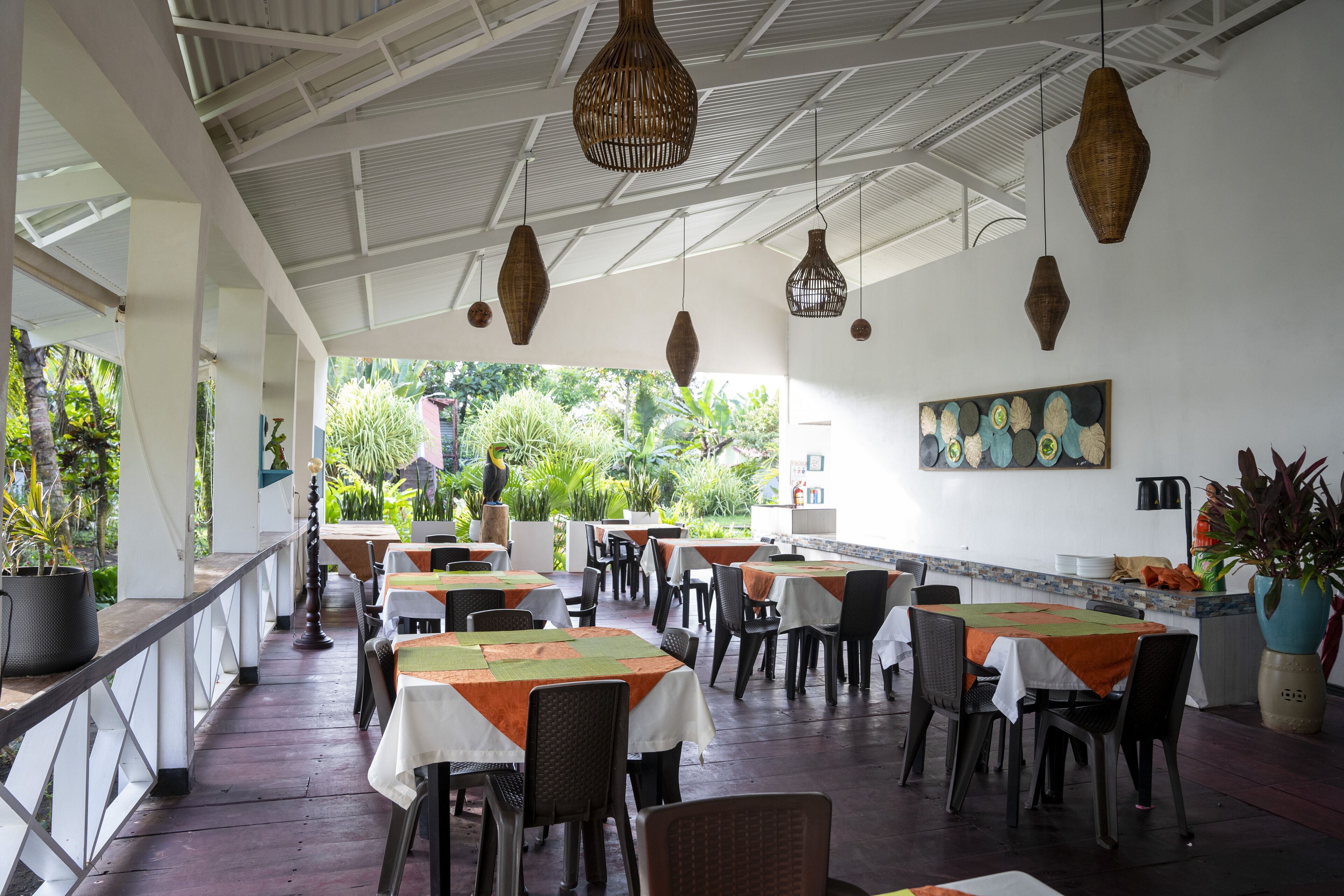 A restaurant with tables covered in white and orange tablecloths, hanging wicker lamps, and a view of greenery outside.