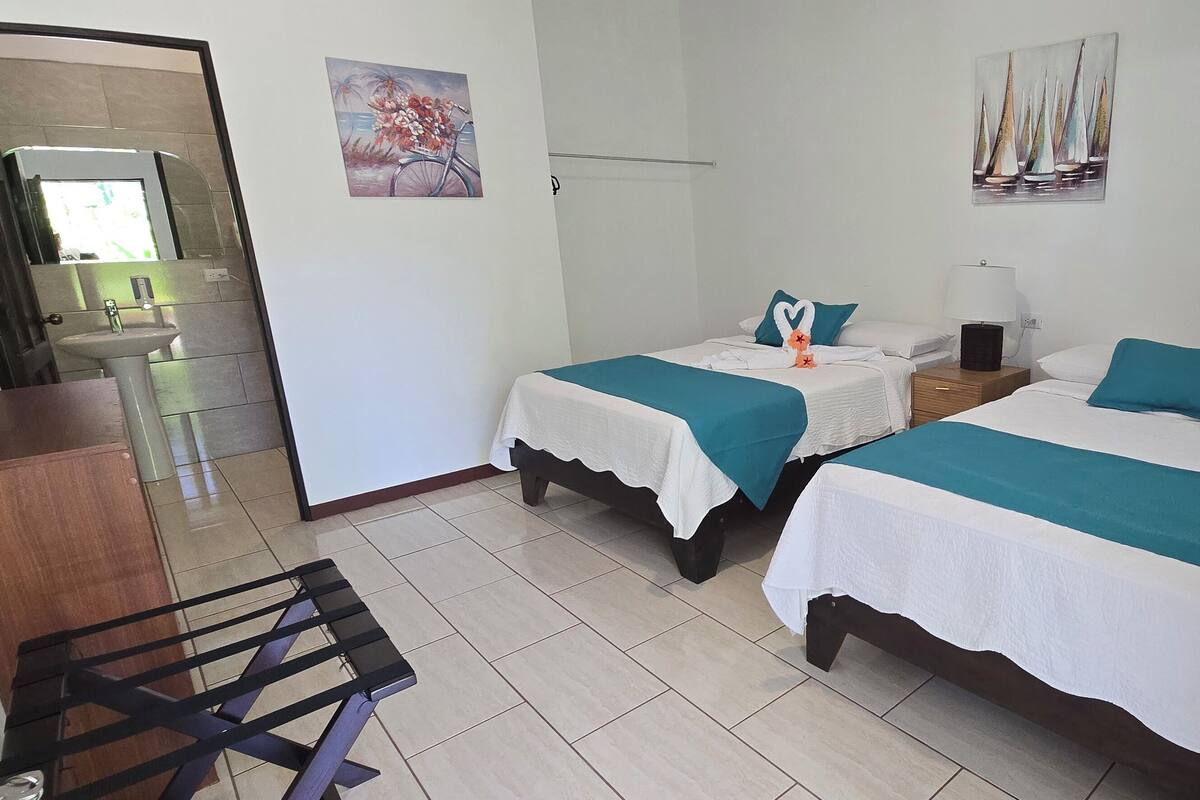 A hotel room with two beds, a bedside table with a lamp, a small sink, and a wooden chair.