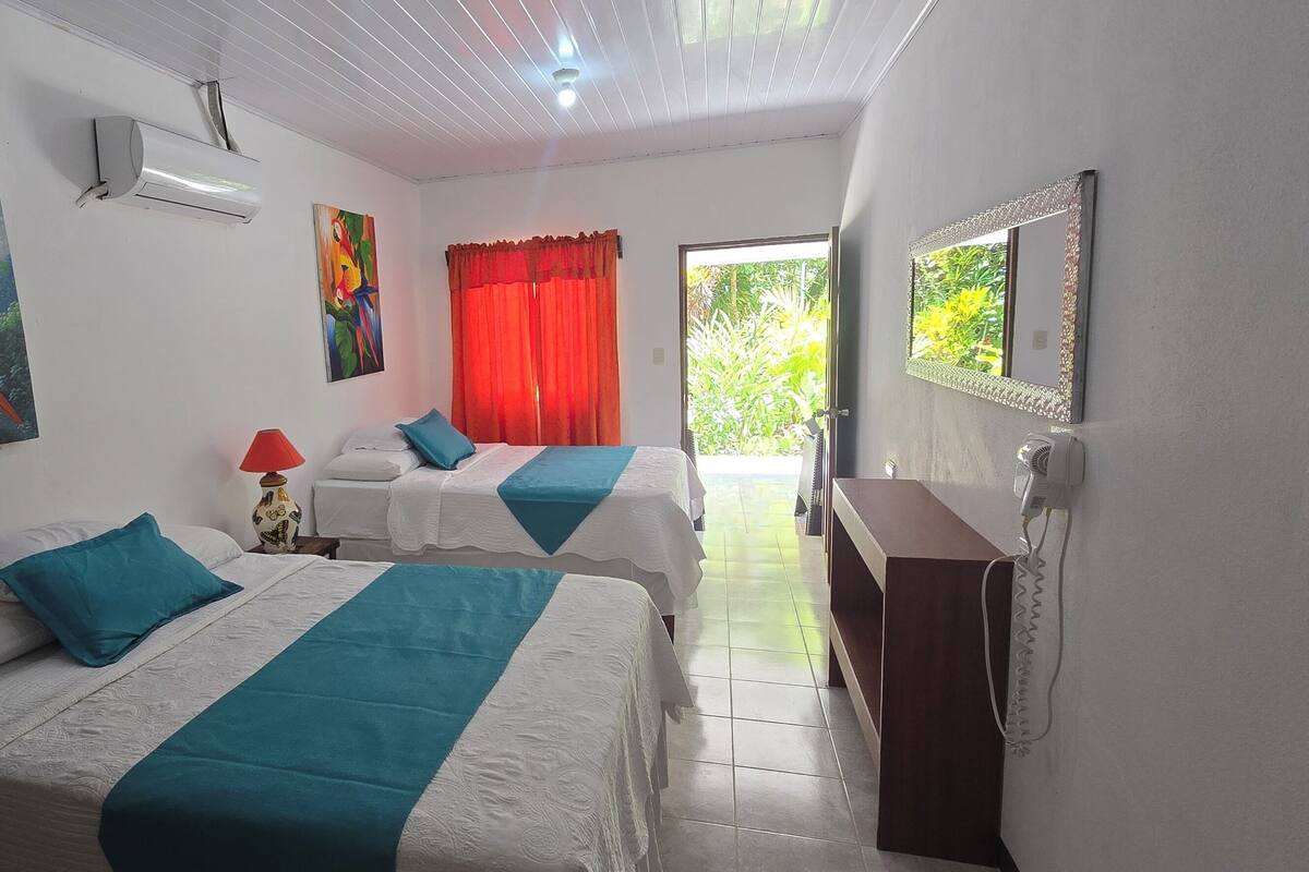 A hotel room with two beds, a red curtain, a painting of parrots, an air conditioner, and a view of greenery outside.