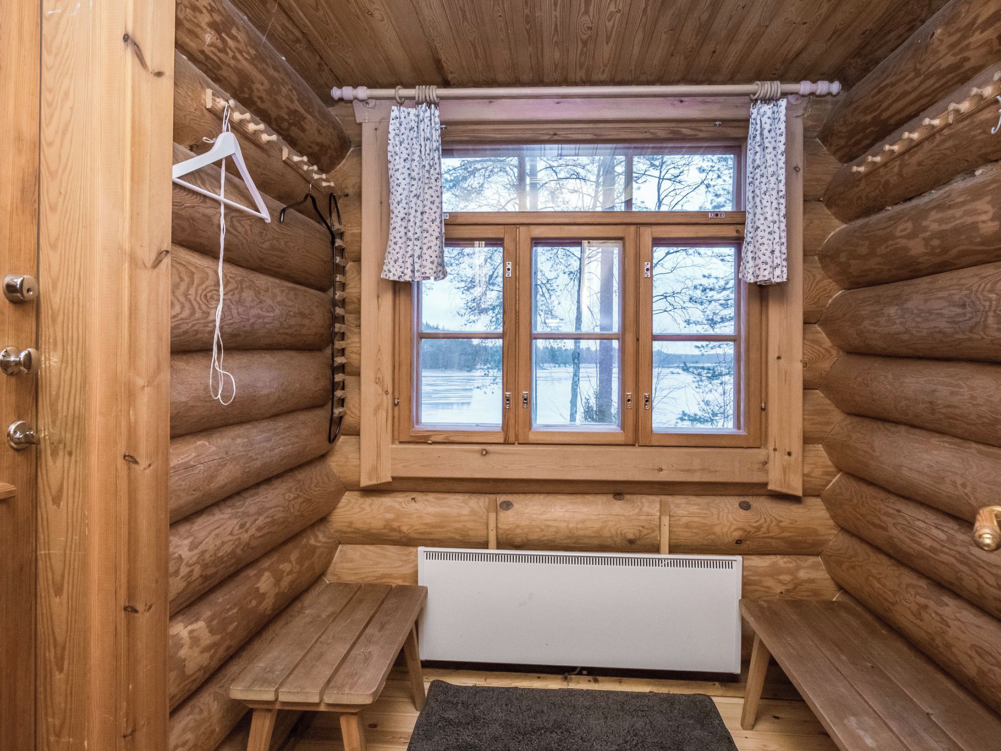 Interno di una baita di legno, con una panca, un termosifone e una finestra con tende.