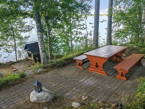 Ein Picknicktisch mit Bänken, ein Grill und eine Laterne auf einem gepflasterten Platz, umgeben von Bäumen und einem Gewässer.